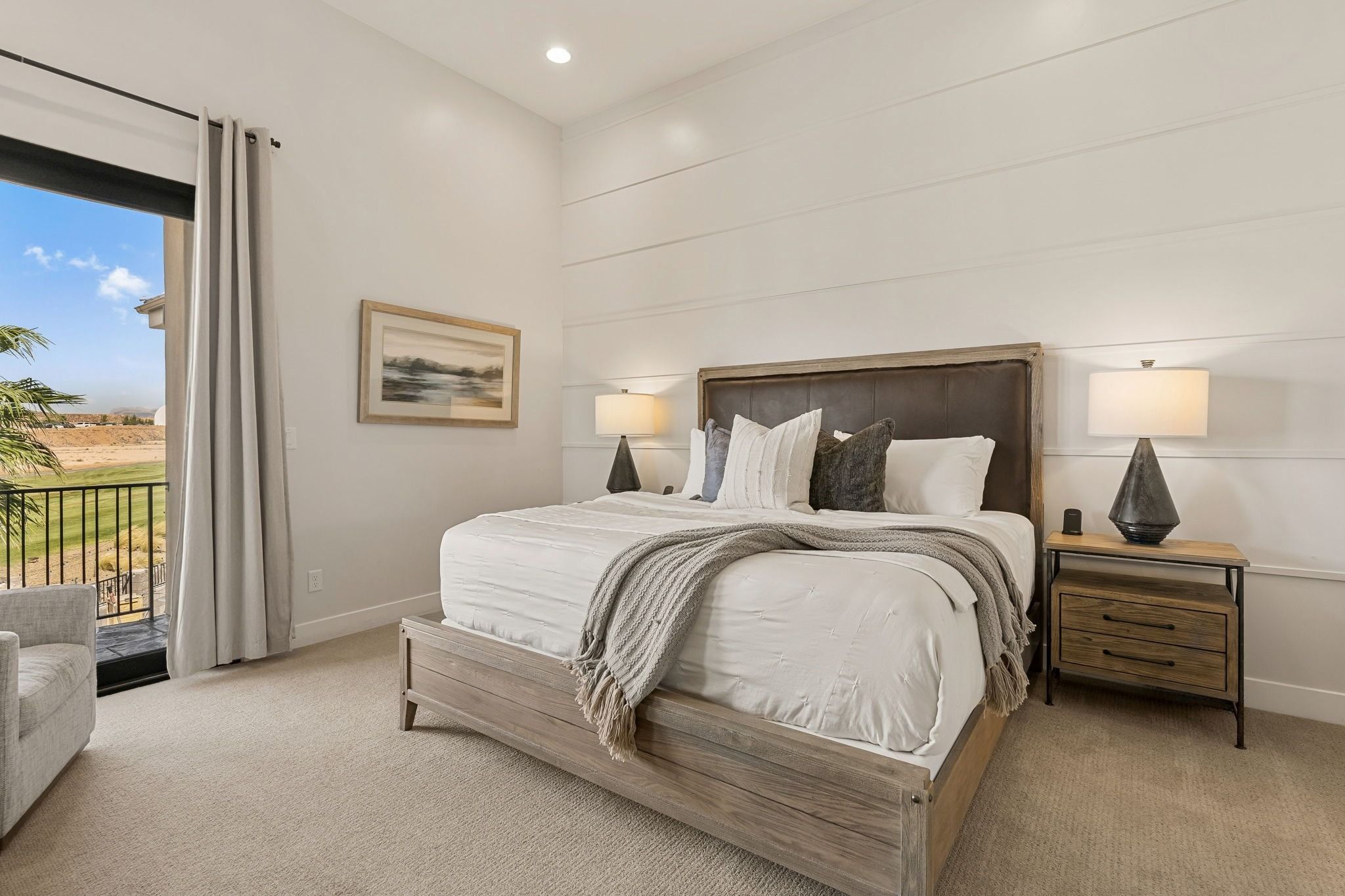 A bedroom with a large bed, two bedside tables, a nightstand, a painting, and a view of a landscape through a sliding door.