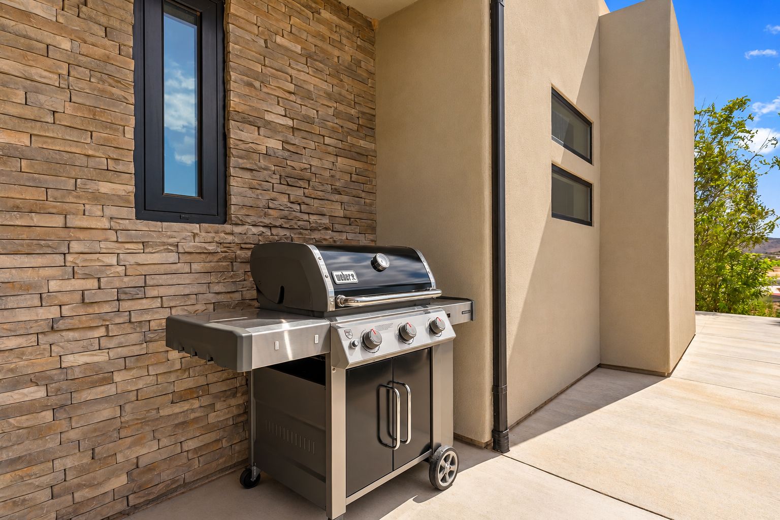 A portable gas grill with four burners and a side cabinet, positioned against a brick wall with a window.