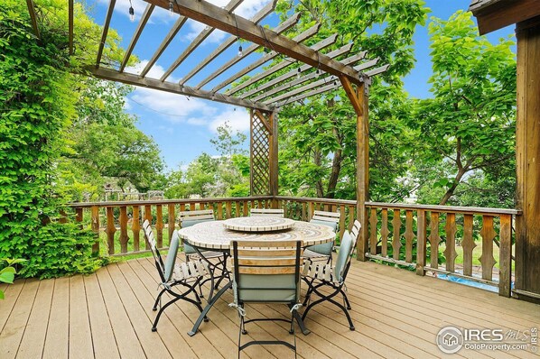 En träaltan med en pergola, ett runt bord med stolar och utsikt över frodig grönska.