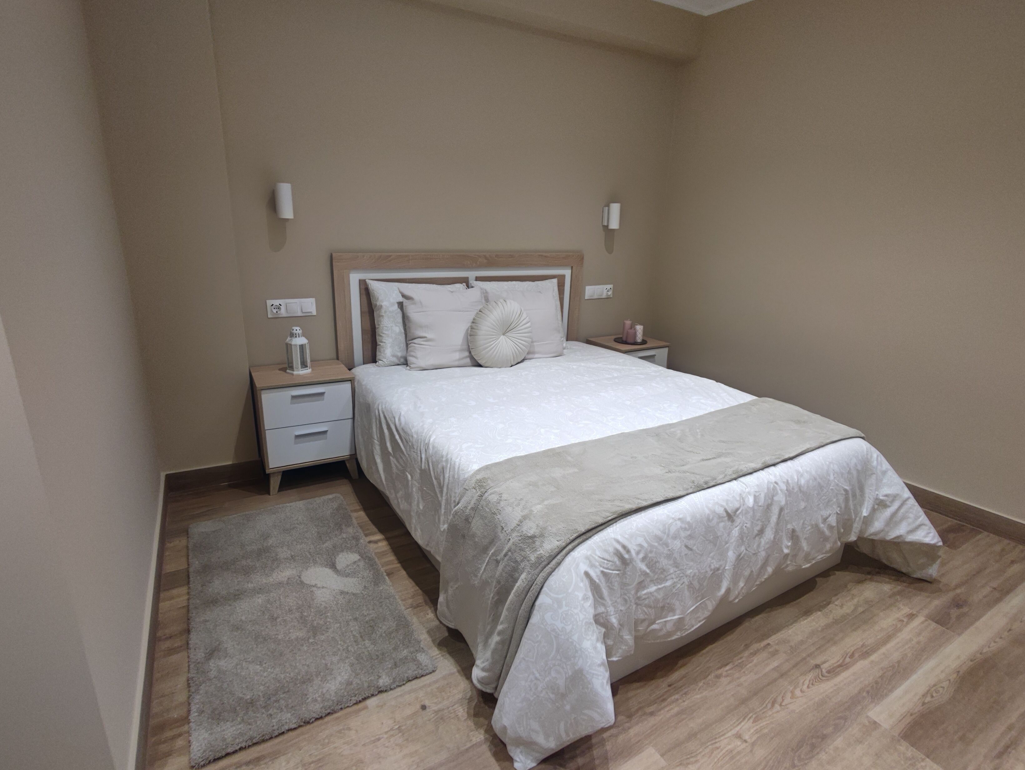 Un dormitorio con cama, mesitas de noche, una alfombra y un aplique de pared.