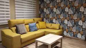 Un sofá amarillo con varios cojines, una mesa de centro de madera y una pared con estampado de hojas.
