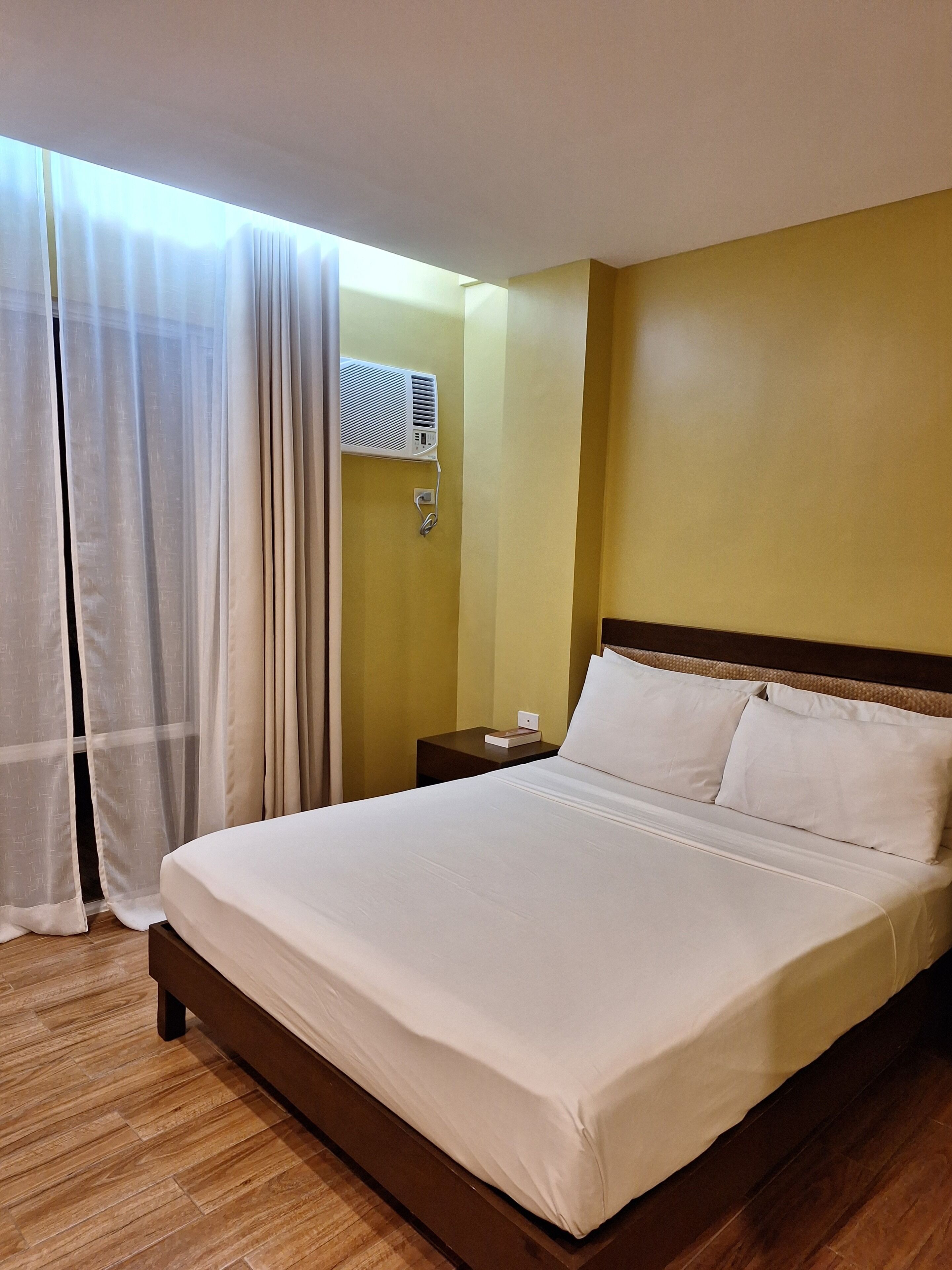 A hotel room with a large bed, a bedside table, a wall-mounted air conditioner, and sheer curtains.
