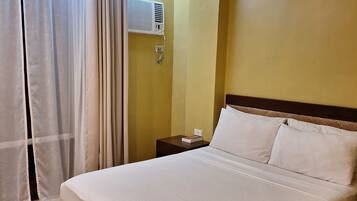 A hotel room with a large bed, a bedside table, a wall-mounted air conditioner, and sheer curtains.