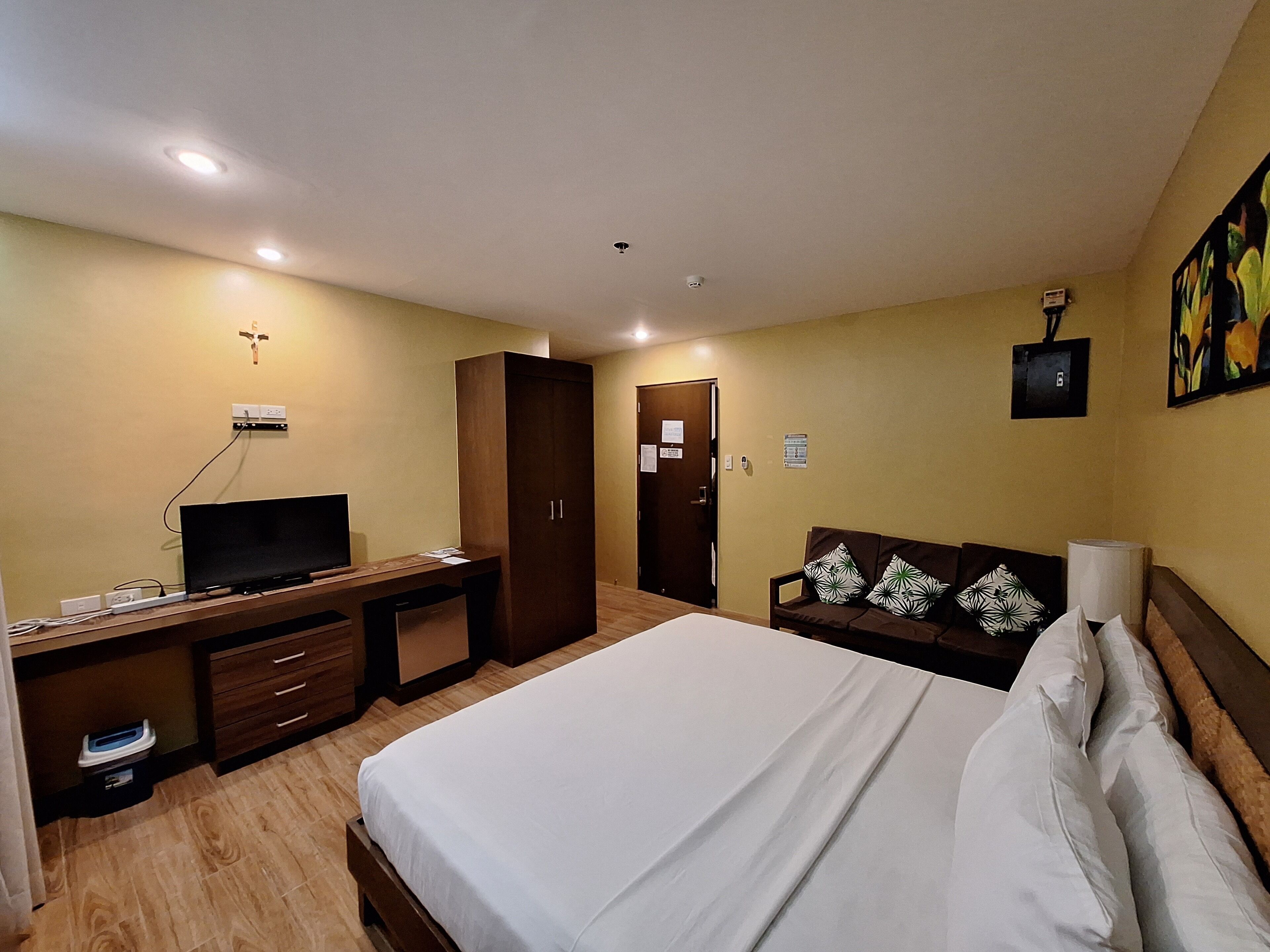 A hotel room with a bed, a desk with a television, a wardrobe, a sofa, and a painting on the wall.