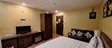 A hotel room with a bed, a desk with a television, a wardrobe, a sofa, and a painting on the wall.