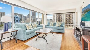 A spacious living room with two sofas, a coffee table, and a TV. Large windows offer a city view.