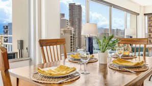 A dining table set with yellow napkins, wine glasses, and a lamp, with a view of tall buildings through large windows.