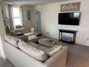 A living room with a beige sofa, a flat-screen TV mounted on the wall, and a small table with a remote control.