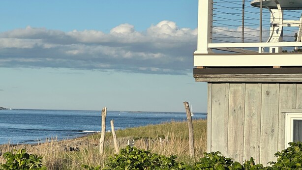 A coastal building with a wooden exterior, a fenced area with vegetation, and a view of the ocean. - Biddeford Pool Beach Condo - Steps to the Sand - 1BR/1BA (Biddeford)