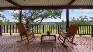 Une terrasse en bois avec deux fauteuils à bascule et une table, offrant une vue panoramique sur un paysage verdoyant, avec des arbres et un ciel dégagé.