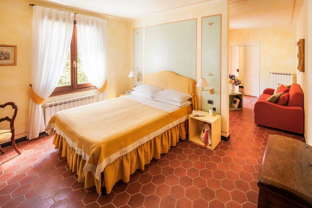 Une chambre d’hôtel avec un grand lit, un canapé rouge, un banc en bois, une fenêtre avec des rideaux et un radiateur.