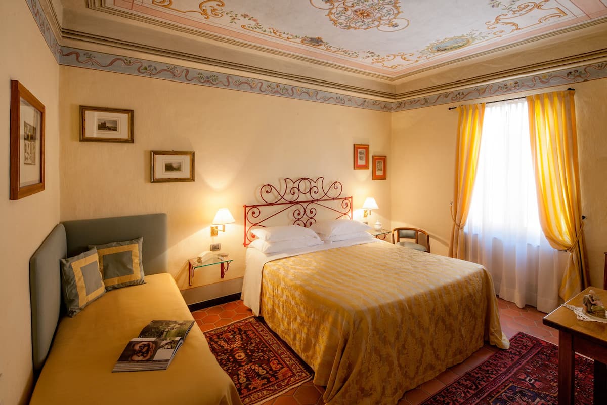 Une chambre d’hôtel avec deux lits, une grande fenêtre avec des rideaux jaunes, un plafond décoratif et des tableaux encadrés accrochés aux murs.