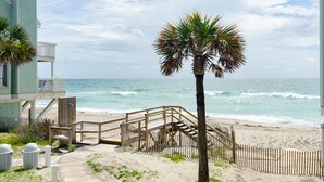 Ein Strand mit einer Holztreppe, die zu einem Gebäude führt, Palmen und Blick auf den Ozean.