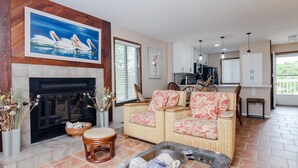 Ein Wohnzimmer mit Kamin, Rattan-Sofa und Essbereich, im Hintergrund ist eine Küche zu sehen.