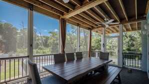 Una terraza cubierta al aire libre con una mesa y sillas de madera, ventanas amplias y vistas al entorno verde.