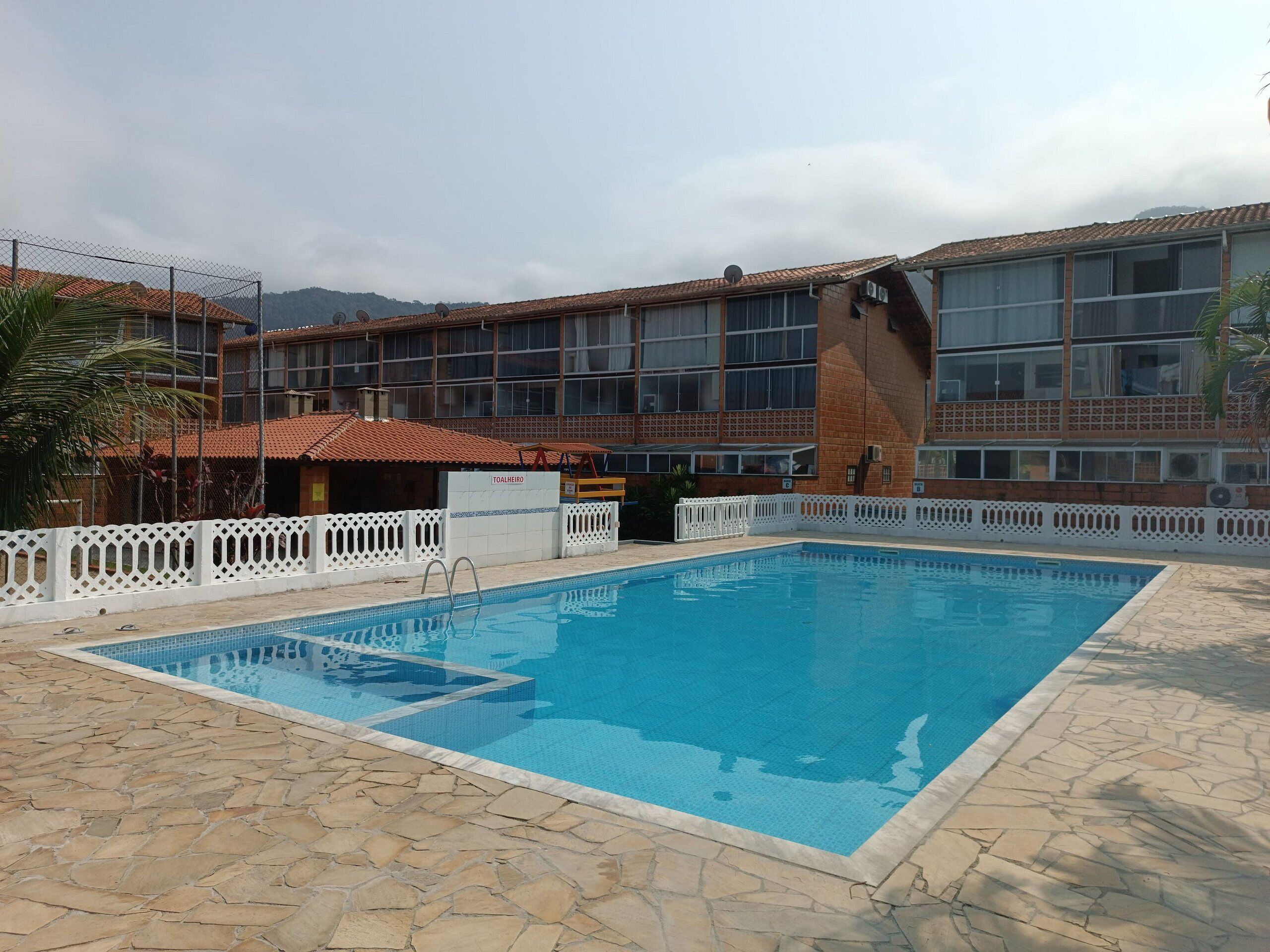 Una piscina con solárium de piedra, cerca una valla blanca y, al fondo, un edificio con tejado de baldosas.