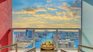 A balcony with a cityscape view, a glass railing, and a wicker basket with bottles and glasses. - Spectacular Studio: Balcony View, Pool, Gym (Hallandale Beach)