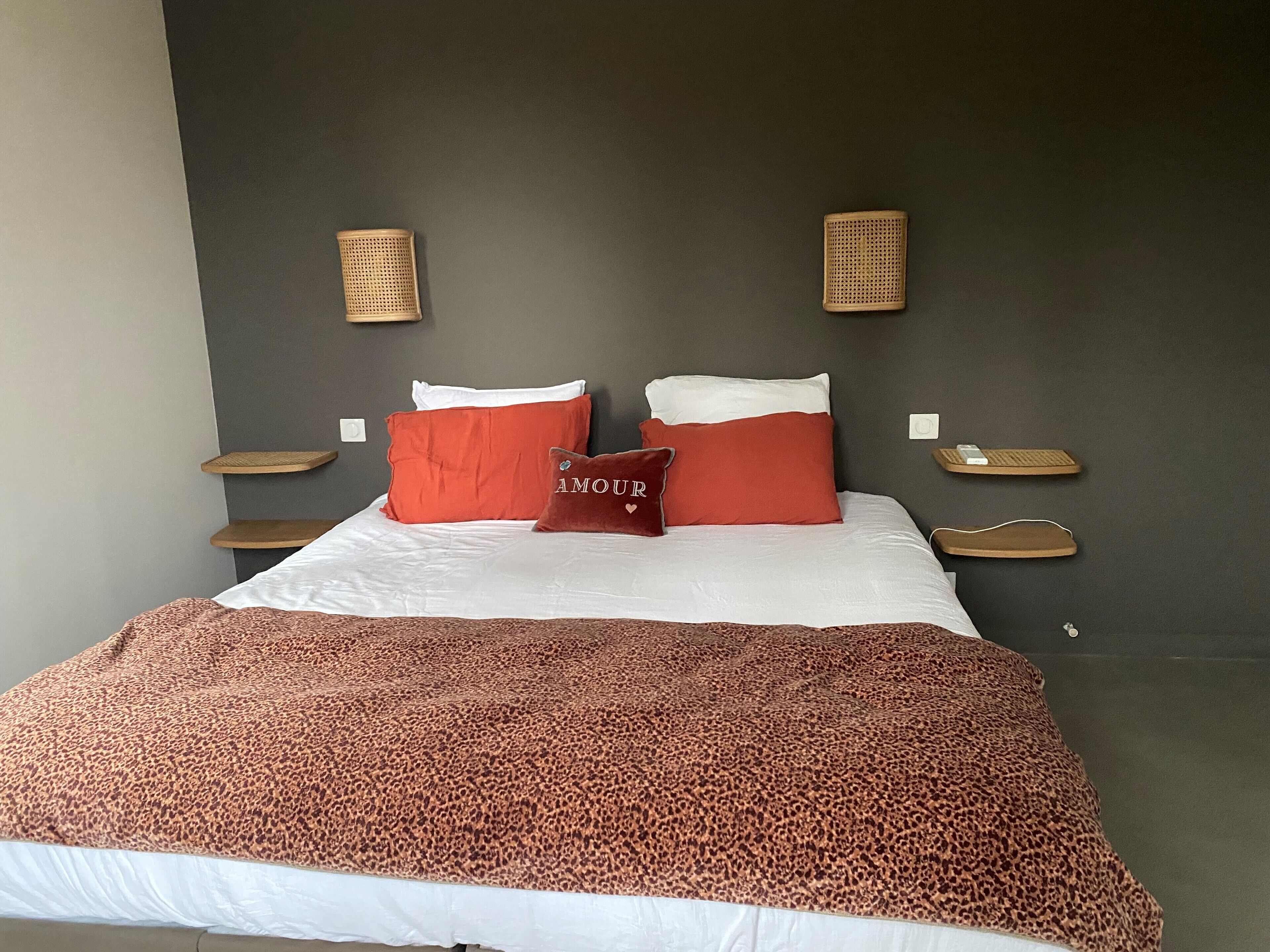 Een bed met wit en rood beddengoed, twee rode kussens, een luipaardprintdeken, en twee wandlampjes met elk een klein tafeltje.