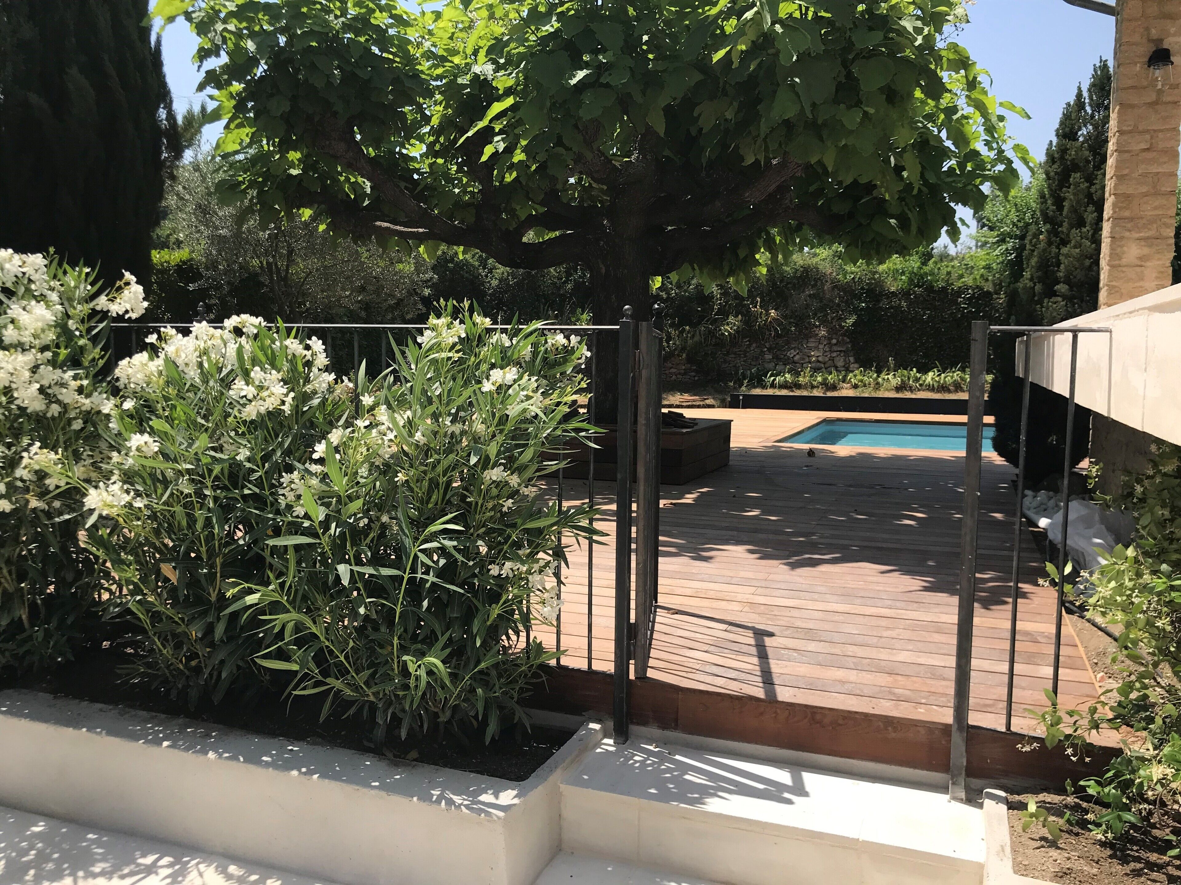 Une terrasse en bois avec une piscine, entourée de verdure et délimitée par une clôture en métal.