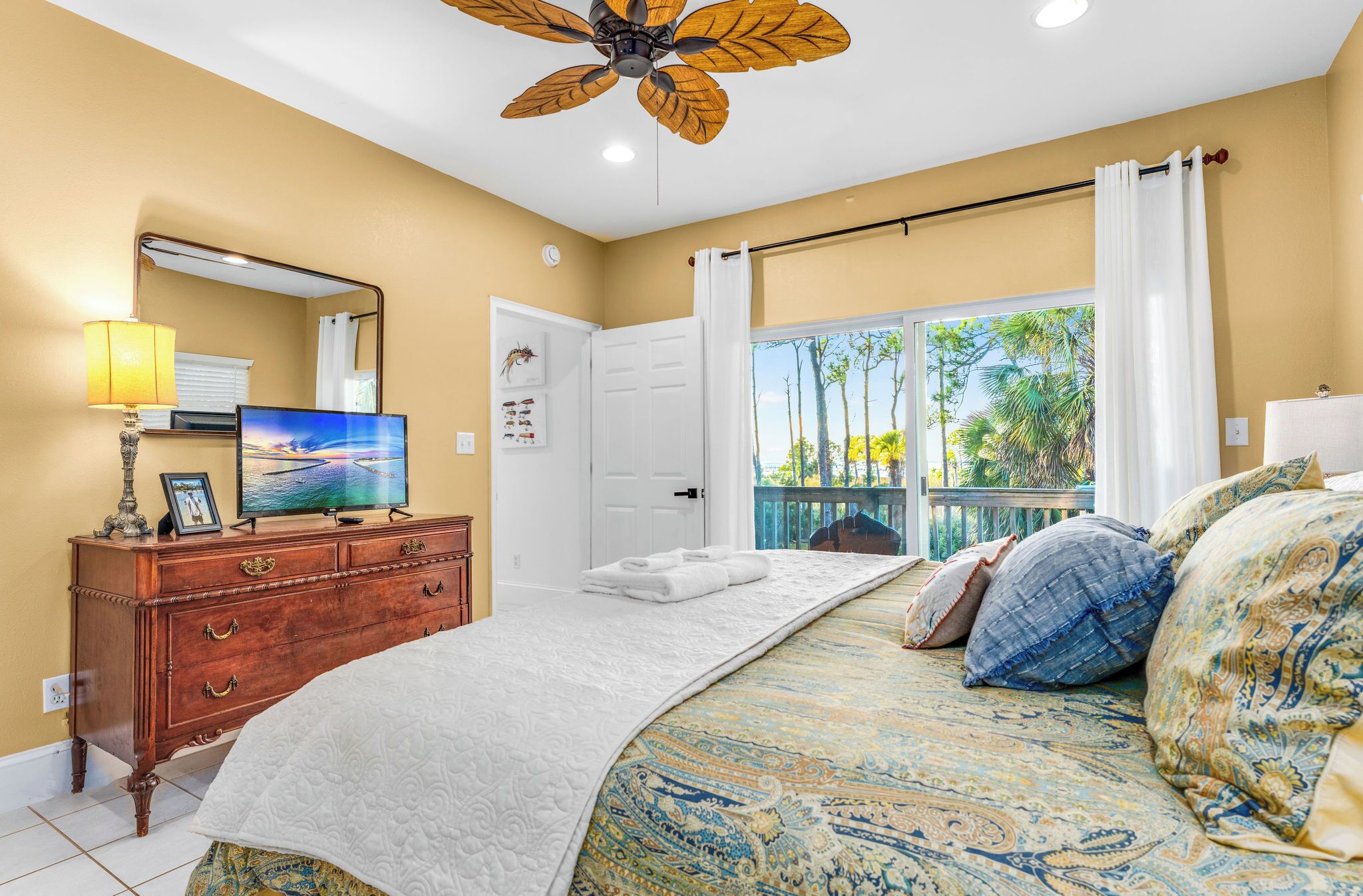 A bedroom with a bed, a dresser, a television, a ceiling fan, a window with curtains, and a door.