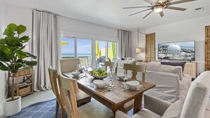 Un'area pranzo con tavolo in legno apparecchiato per quattro persone, una ventola a soffitto e vista sulla spiaggia attraverso ampie finestre.