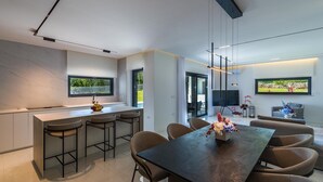 A modern kitchen with a central island, bar stools, a dining table with chairs, and a flat-screen TV mounted on the wall. - Villa Valinka Design (Golas)