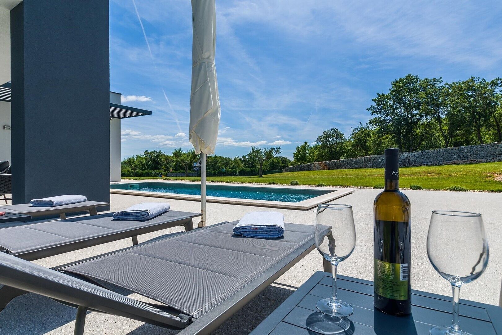 Ein Poolbereich mit Liegestühlen, einer Weinflasche und einem Glas auf einem Tisch, mit Blick auf einen Swimmingpool und Grünflächen.