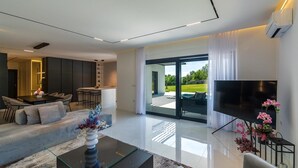 A modern living room with a sofa, coffee table, and TV. There is a dining area with chairs and a table, and a kitchen in the background. - Villa Valinka Design (Golas)