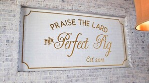 Un letrero en la pared dice "PRAISE THE LORD THE PERFECT FIg Est. 2012" sobre un muro de ladrillos blancos.