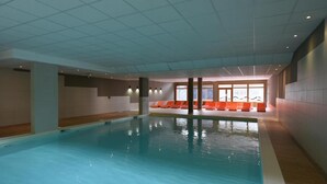An indoor swimming pool with clear water, surrounded by orange lounge chairs and a tiled floor.