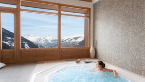 A person relaxing in a hot tub with a mountain view through large windows.