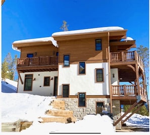Eine zweistöckige Chalet mit Stein Fundament, Holz Balkonen und einer Skipiste im Hintergrund.