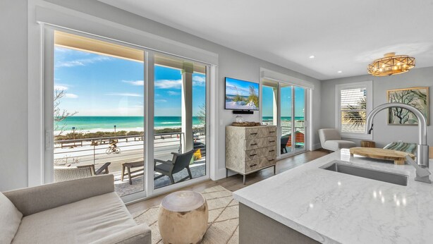 A modern living room with a large window overlooking a beach, a flat-screen TV, and a kitchen area with a marble countertop. - Rooftop deck with Gulf views - Villa Rayo Del Sol (Bradenton Beach)