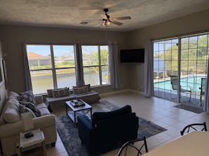 A spacious living room with a sofa, armchairs, a coffee table, and a TV mounted on the wall. Large windows offer views of a swimming pool and greenery. - Blue Oasis 3 Bedroom Home by RedAwning (Cape Coral)