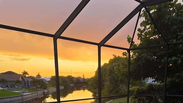 An indoor swimming pool with a view of a sunset over a body of water, surrounded by greenery. - Blue Oasis 3 Bedroom Home by RedAwning (Cape Coral)