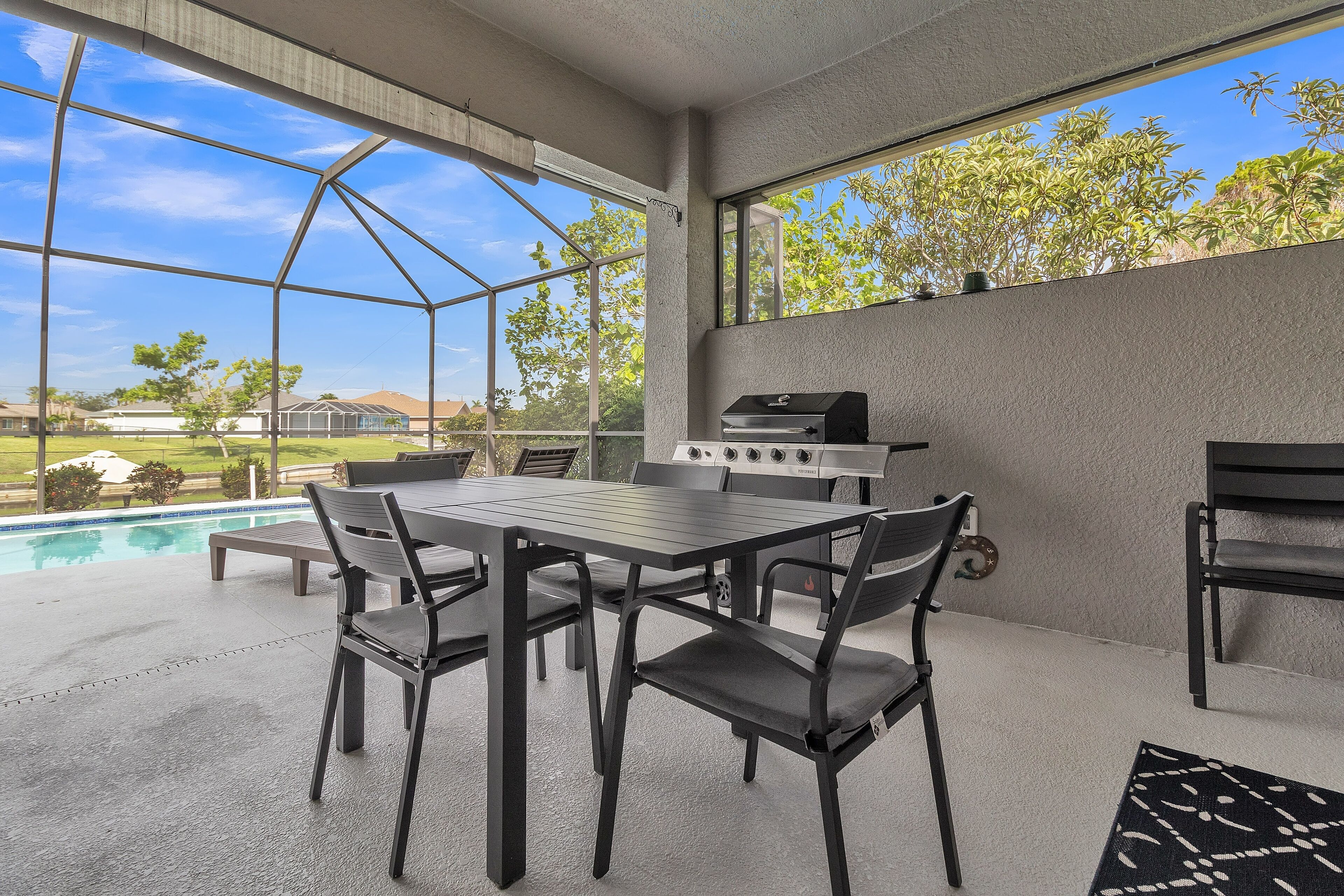 Patio esterno con tavolo e sedie, un barbecue e vista su piscina e vegetazione.