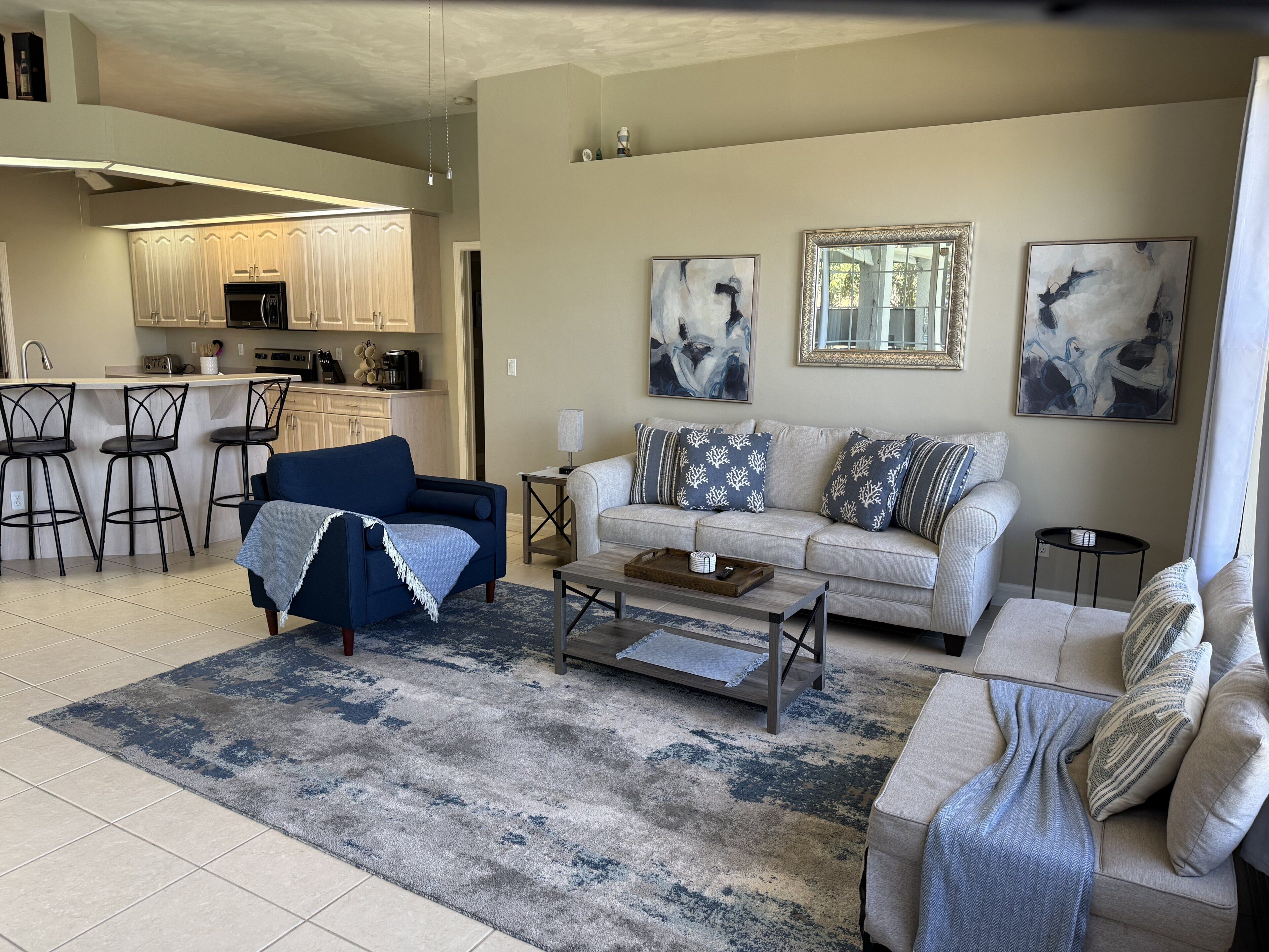 A modern living room with a sofa, coffee table, and chairs, a kitchen in the background, and abstract art on the walls.