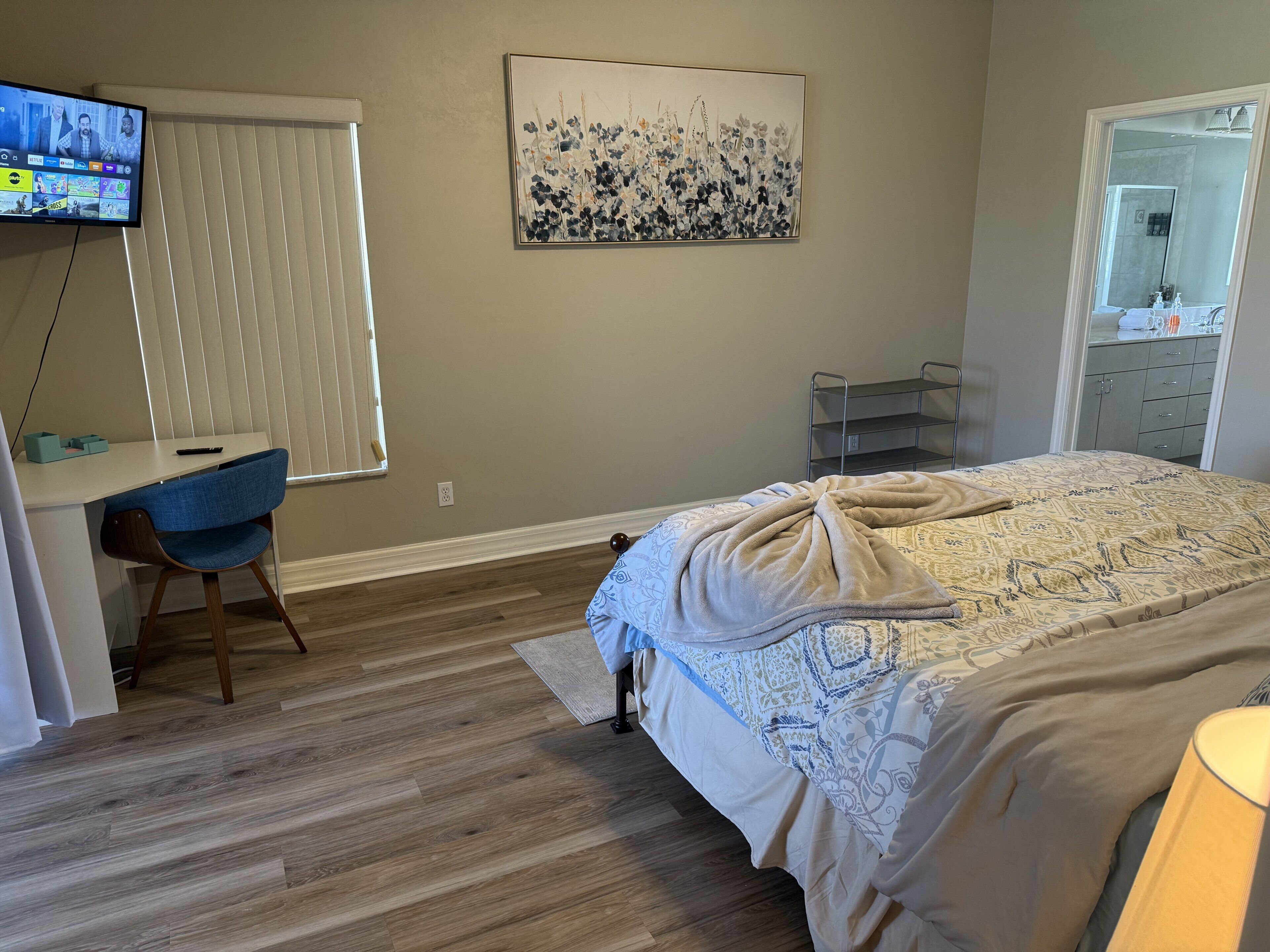 A bedroom with a bed, a desk with a chair, a TV, a painting, and a bathroom visible through an open door.