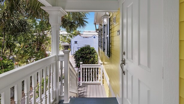 Un escalier étroit avec des rampes blanches mène à une porte jaune, qui est entourée de verdure et offre une vue sur un bâtiment bleu.