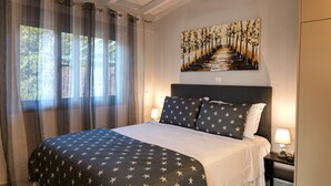 A bedroom with a bed, two nightstands, a ceiling fan, a window with sheer curtains, and a wall-mounted painting. - Gems Apartments (Corfu)