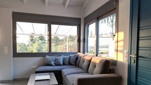 A modern living room with a sectional sofa, a small table, and large windows offering a view of greenery outside. - Gems Apartments (Corfu)