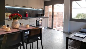 A modern kitchen with a dining area, featuring a large window, a vase of red flowers, and a coffee table. - Gems Apartments (Corfu)