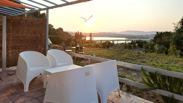 A covered outdoor seating area with white plastic chairs and a table, overlooking a scenic landscape with a body of water and hills in the distance. - Gems Apartments (Corfu)