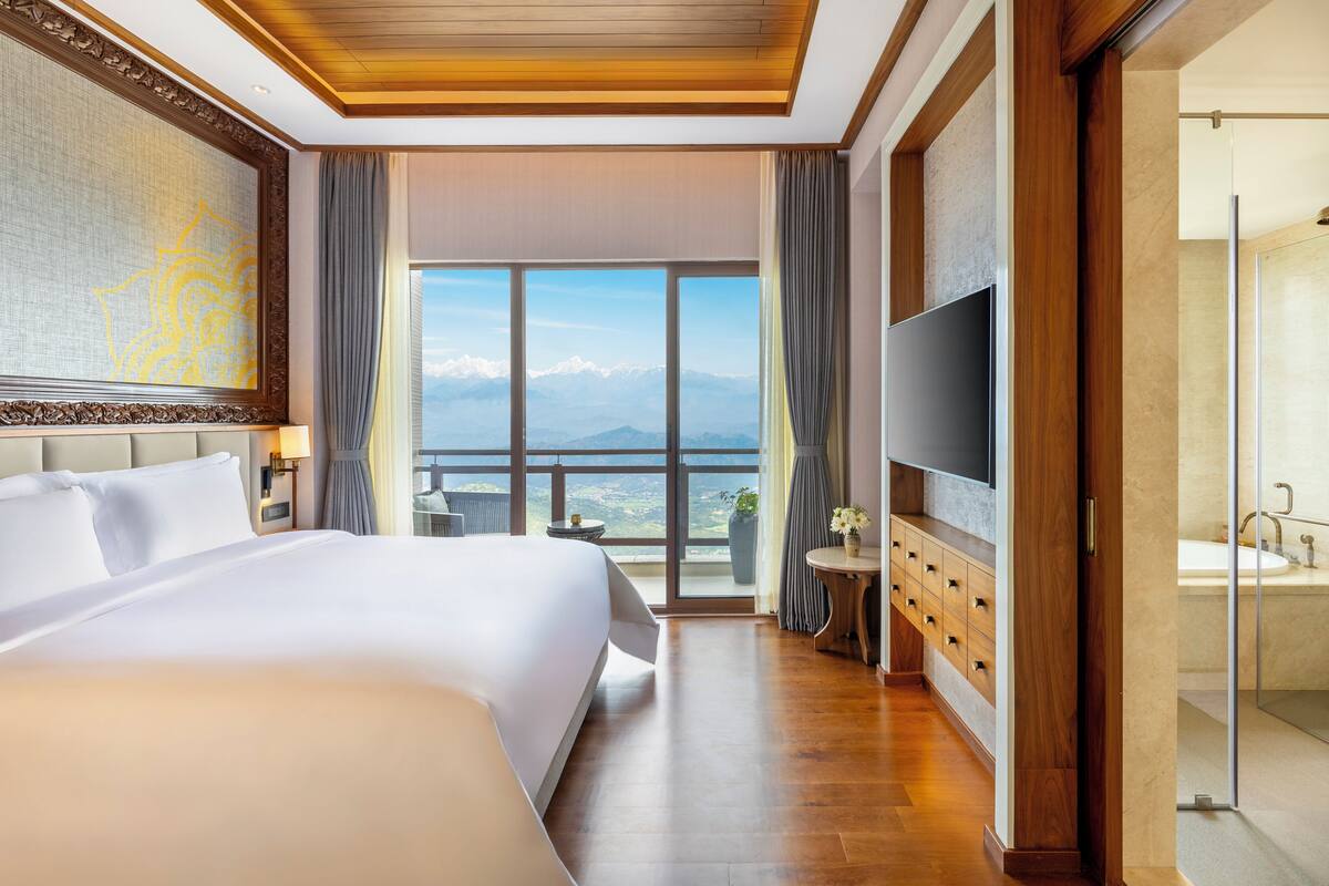 A hotel room with a large bed, a TV, a bathroom with a bathtub, and a view of mountains through the window.