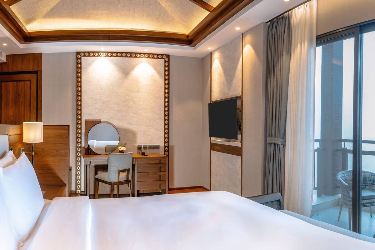 A hotel room with a large bed, a desk with a chair, a mirror, a TV, and a balcony with a view.