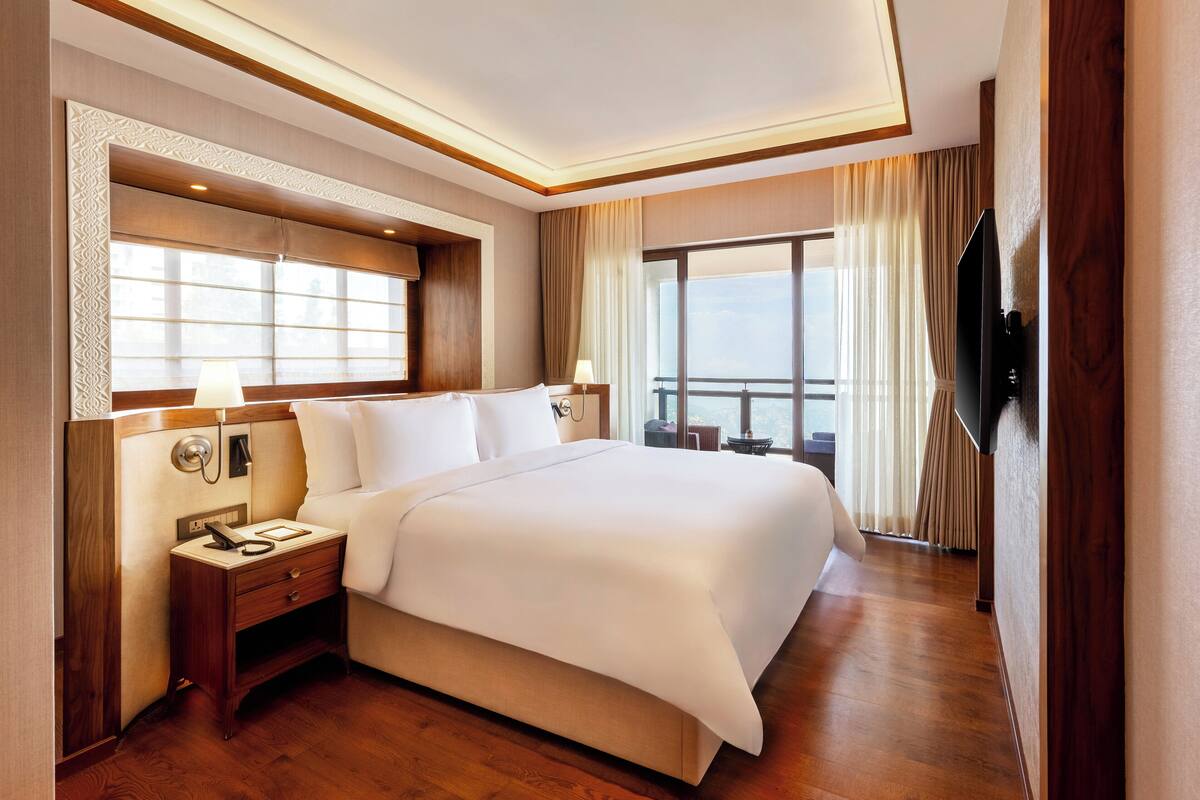 A spacious hotel room with a large bed, a flat-screen TV mounted on the wall, and a balcony with curtains.