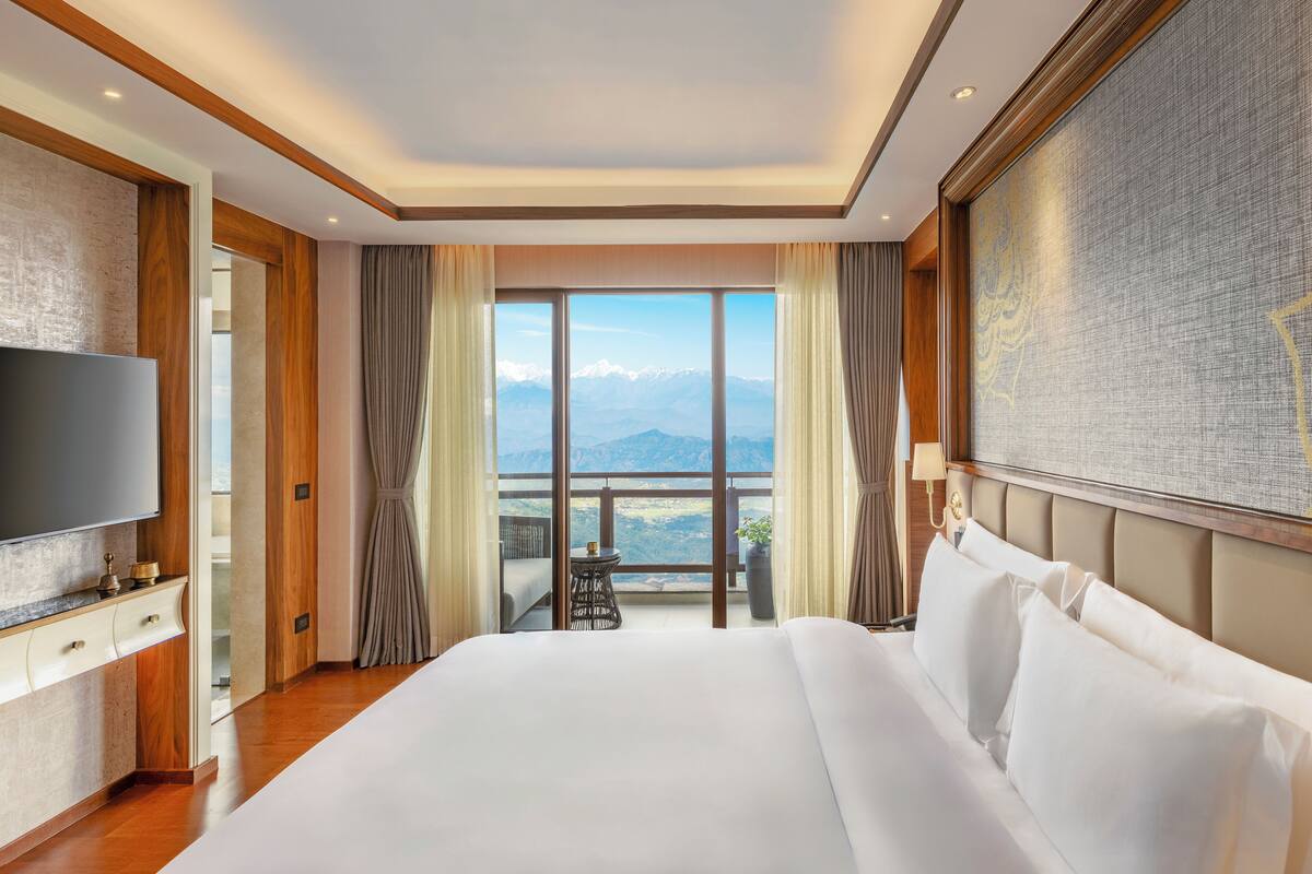 A hotel room with a large bed, a flat-screen TV, a balcony with a mountain view, and a large painting on the wall.
