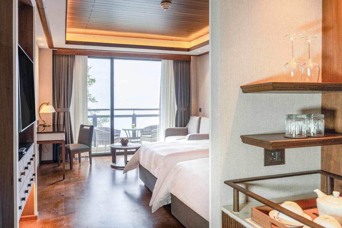 A hotel room with a large bed, a desk with a lamp, a chair, a small table with a bowl of fruit, and a balcony with a view.