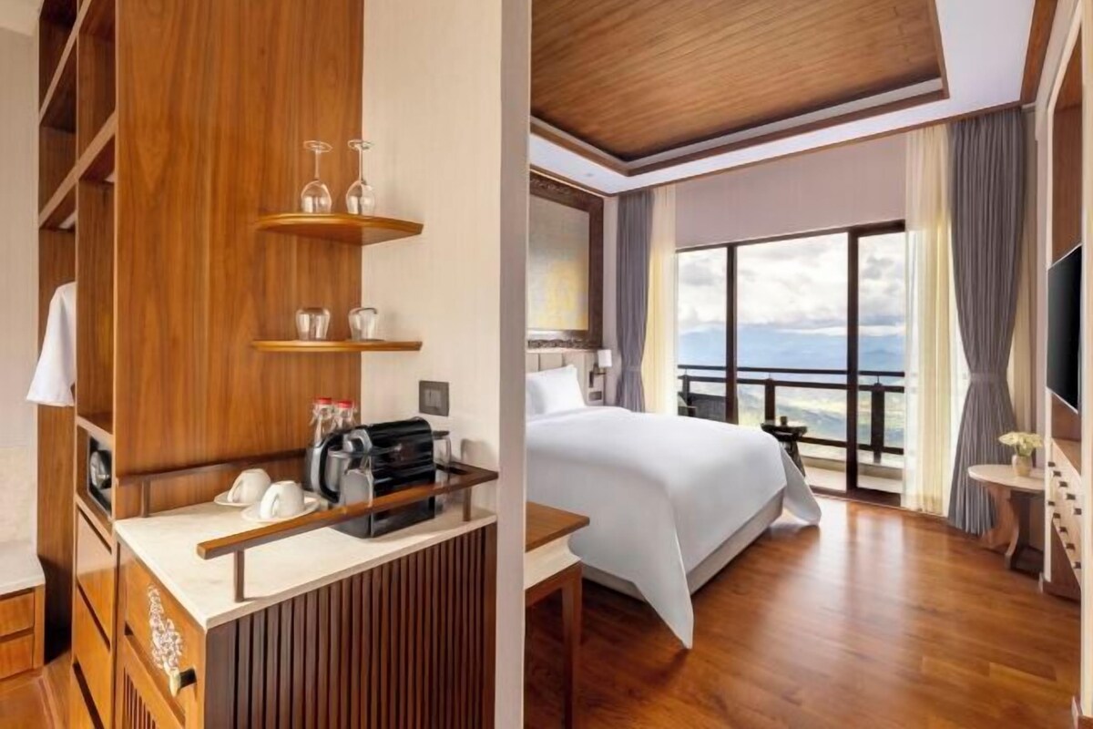 Une chambre d’hôtel avec un bureau en bois, un lit, une grande fenêtre donnant sur un beau paysage et un balcon.