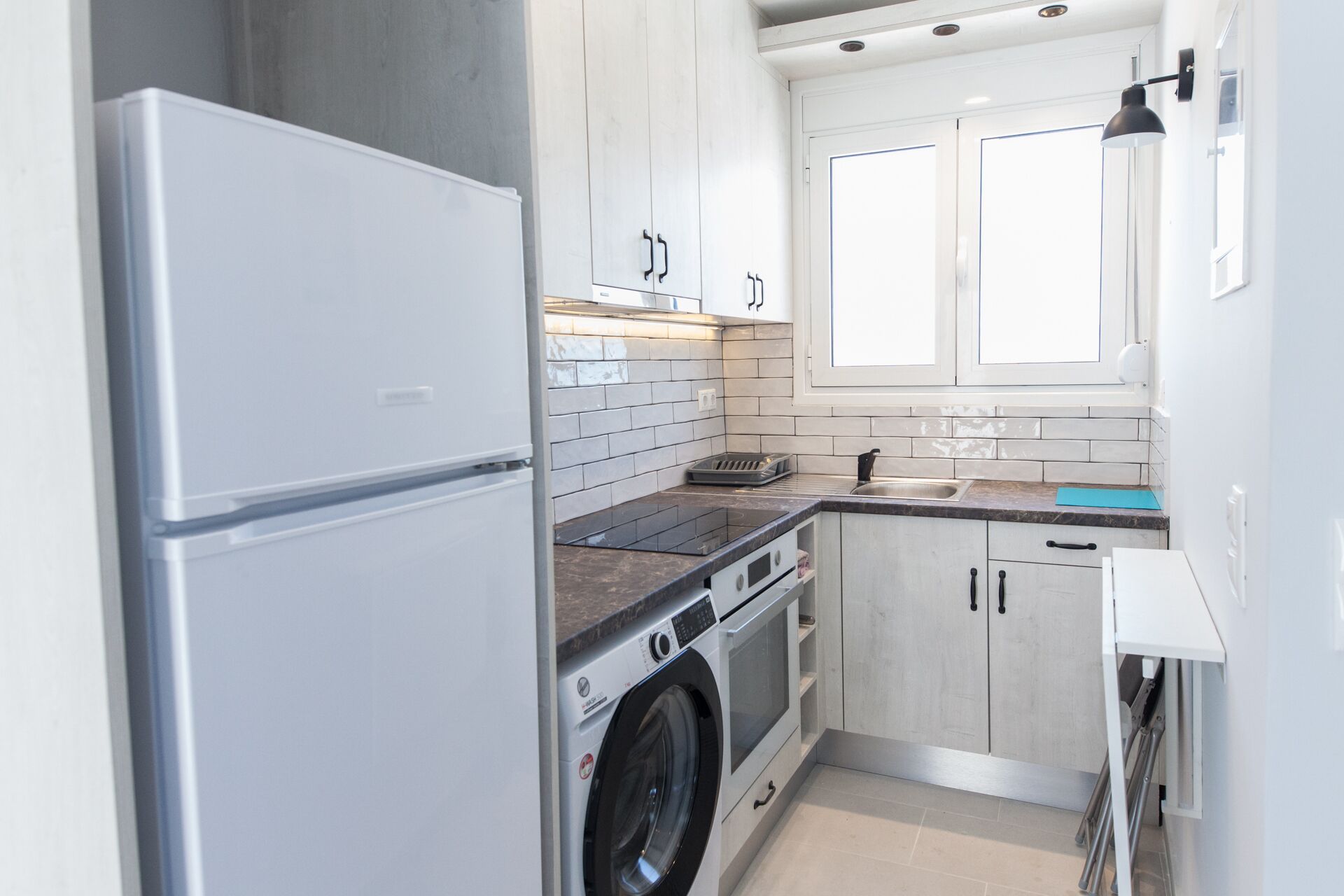 A modern kitchen with white cabinets, a stainless steel refrigerator, a built-in oven, a washing machine, and a window with blinds.